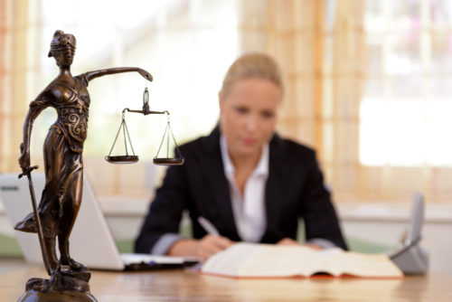 A woman in business attire works with a laptop, book, pen and paper in front of her. A statue of Lady Justice holding scales and sword sits on the desk.