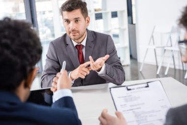 Man in suit counting on fingers during an interview while one professional points and the other professional takes notes.