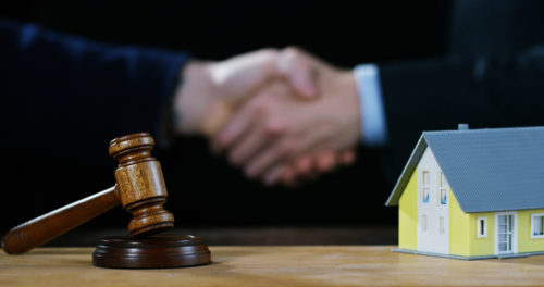 A gavel and house rest in the foreground while a handshake in the background finalizes a bid at an auction.