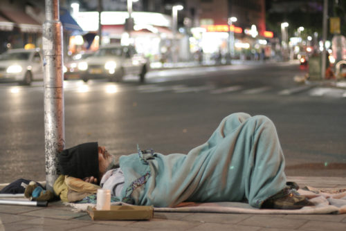 Homeless man sleeping on a street corner with a single blanket covering him.