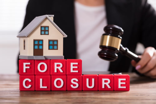 A woman holds a gavel at a foreclosure property auction, with a house on top of blocks spelling out “foreclosure.”