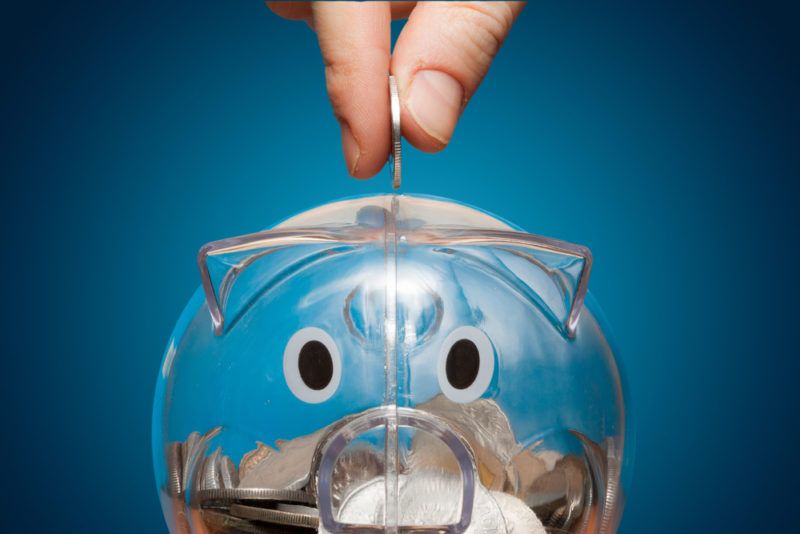A hand deposits a quarter in a clear piggy bank.