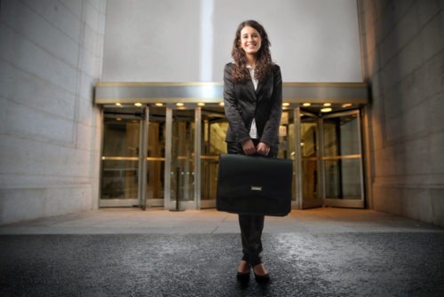 A woman wearing professional attire holding a briefcase stands outside an office building.