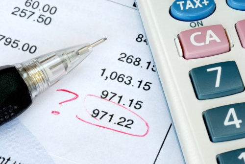 A bank statement with a list of charges. A charge for $971.22 is circled in red pen with a question mark. A calculator and pen sit on the paper statement.