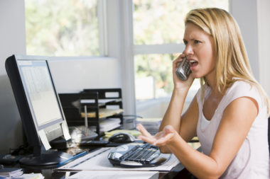 A woman sitting at a desktop computer on the phone visibly upset.