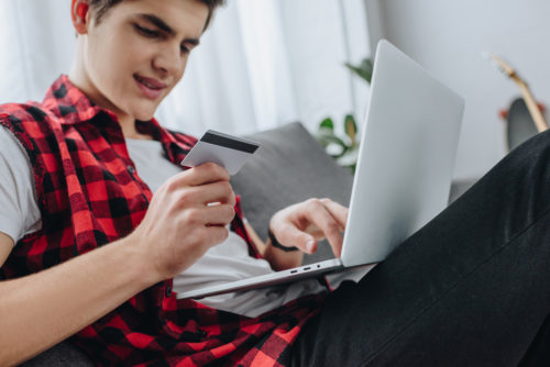 Teenager shopping online with a laptop and credit card. He is wearing a red checkered shirt and dark jeans.