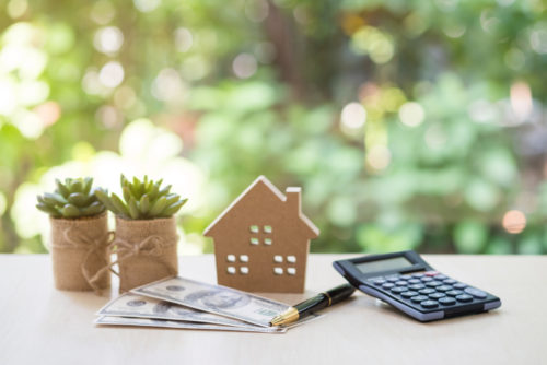 Concept photo showing home refinance. Two succulents, model of a house, three 100 dollar bills, a pen, and a calculator on a table.