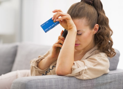 A consternated woman on the phone, holding a credit card, rests her forehead against her hand.