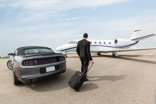 A man in a business suit exists a charcoal Mustang convertible car and is walking towards a small private jet on a runway.