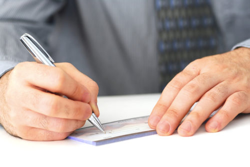 A man in a business suit sits at a table, writing out a check with a silver pen.
