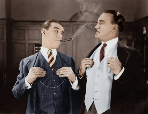 Two men in finely tailored suites smoking cigars and looking at each other.