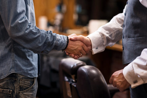 A close up of two men shaking hands; one man is wearing casual jeans and a shirt, while the other has an expensive looking button up vest, black slacks, and a white collared shirt.