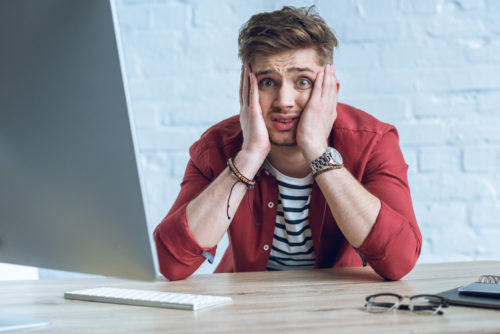 A stressed man holds his head in his hands while looking at the camera, past a computer monitor.