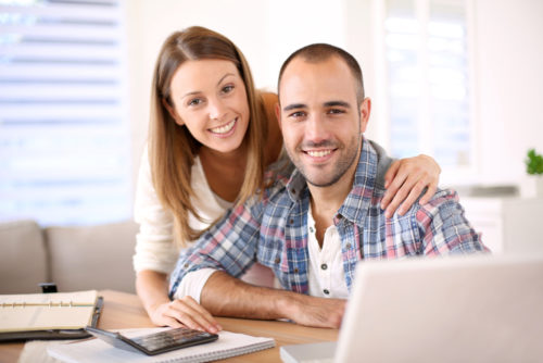 A man sits at a table with a woman leaning over his shoulder. A laptop, calculator, and notepad sit on the table in front of them.