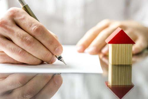 Close up of a man’s hands with a pen signing closing agreement with a small wooden house model next to it