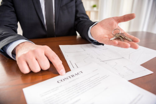 Close up of a man in a suit holding out a set of two keys with one hand and pointing at a “contract” on the desk with his other hand.