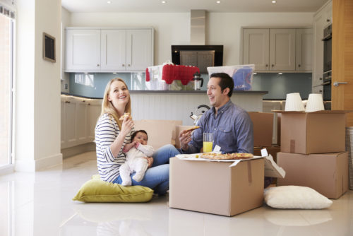 A young couple with a baby are sitting on the floor of the kitchen in their new home, enjoying pizza and smiling.