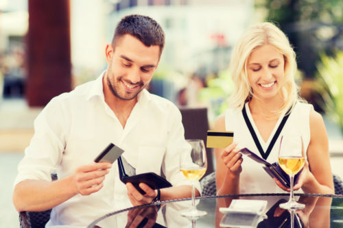 A couple, drinking wine outside at a restaurant, each pull out a credit card to pay for a date.