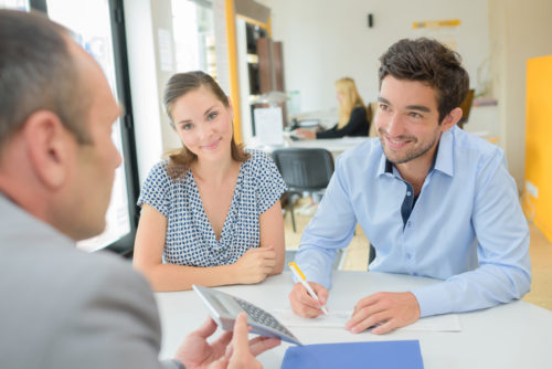 A smiling young couple signing papers with a loan officer holding a calculator in a bank.