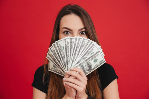 A woman holds hundred-dollar bills in front of her face, against a red background.