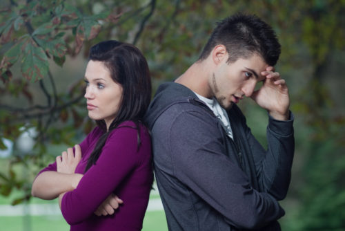 A young couple with relationship problems stands back to back, the woman crossing her arms while the man holds his head in a hand.