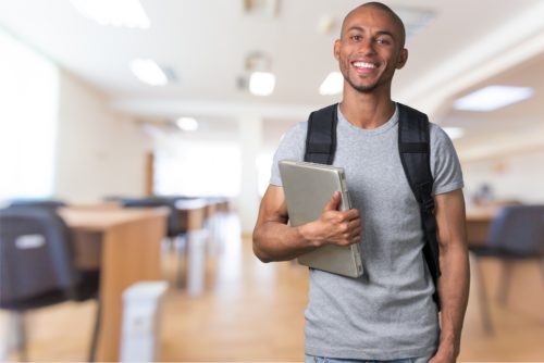 A male student wearing a backpack holds a laptop and stands in a classroom