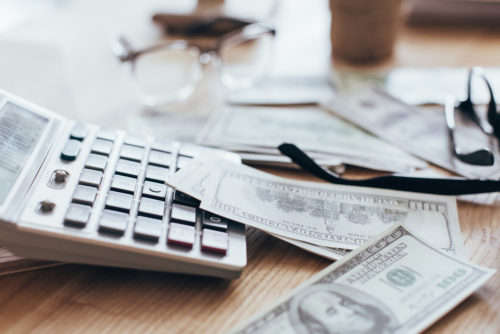 Several $100 bills laying on top of a calculator next to a pair of glasses