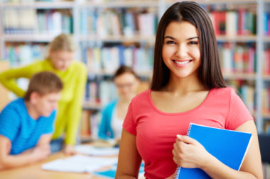 Girl in college library, holding a blue spiral notebook, with her group mates in background, out of focus.