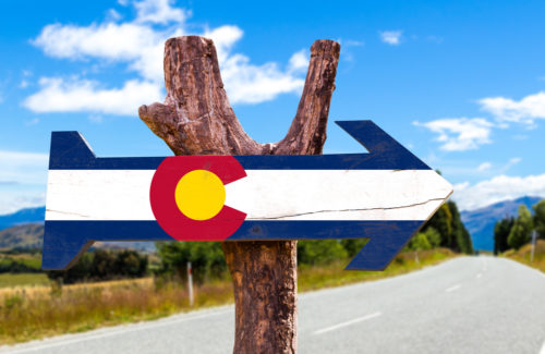 A sign shaped like an arrow with the Colorado state flag printed on it hanging on a tree branch.