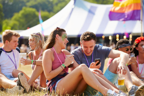 Millennials sit on the grass, drinking and looking at their phones, while enjoying an outdoor music festival.