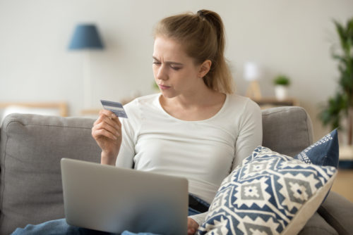 A confused woman sits on a couch with a laptop and holds a credit card