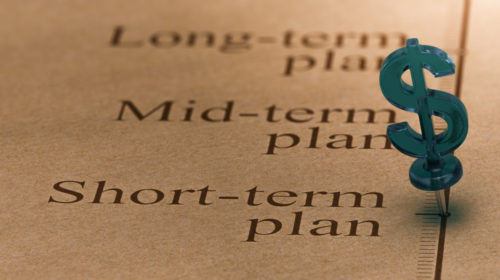 A close-up photo of a dollar-shaped pushpin placed on a timeline next to the words “short-term plan.”
