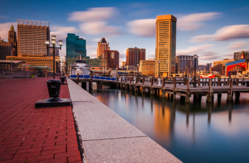 A long-exposure photograph of the Baltimore, Maryland skyline, against a blue sky dotted with clouds.
