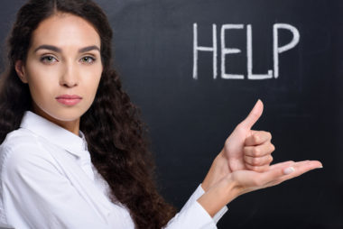 An image of a woman gesturing sign language for the word "help" with the word written behind her in chalk.