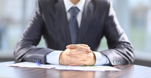 Male in a suit sits at a table with his hands folded over paperwork.