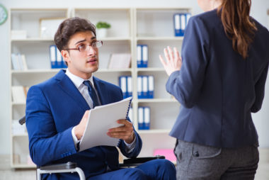 An image of a man in a wheelchair writing on a notepad and having a discussion with a womsn.