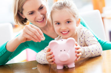 A kid holds a piggy bank as her mother is reaching over her, putting money into it for savings.