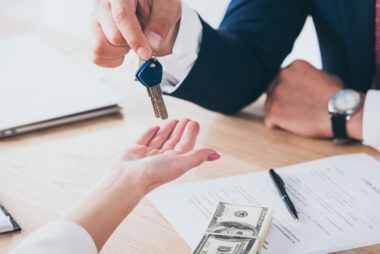 Two people sitting at a table with car loan papers and money sitting on top of it. One person is handing the other a set of keys to a car.