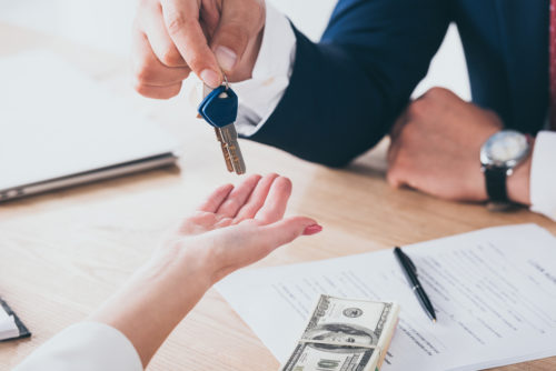 Two people sitting at a table with car loan papers and money sitting on top of it. One person is handing the other a set of keys to a car.