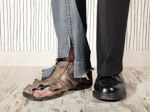 A man’s legs are shown below the knee, with torn jeans and a worn-through work shoe on one leg/foot, and black slacks and a dress shoe on the other leg/foot in order to juxtapose poverty and wealth.