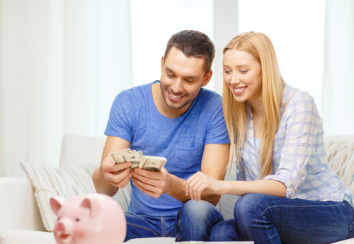 A smiling couple sitting on a couch counting money with a piggy bank in front of them.