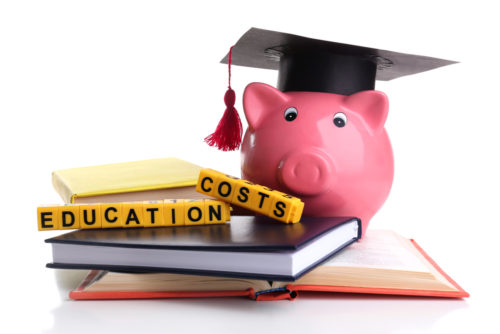 A piggy bank with a graduation cap on sitting atop some school books, next to some word blocks displaying the words "education costs."