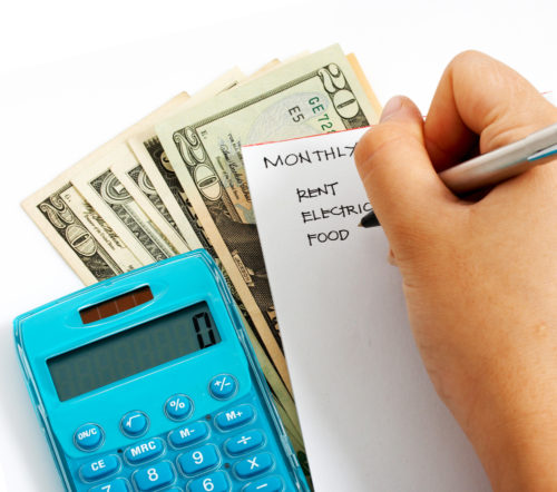 A hand writes out a budget on top of some money and next to a calculator.