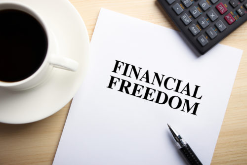 A coffee cup sits on a desk next to a calculator and a paper that has the words "financial freedom" printed on it.