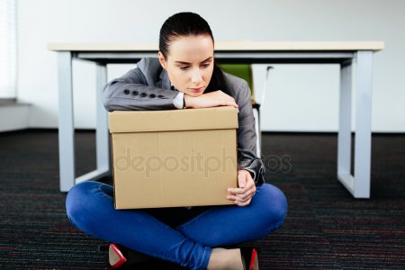 Sad woman holding a box and sitting on the floor in front of her empty office desk.