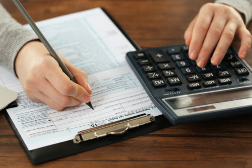 A man sits at a desk typing on a calculator on one hand and writing on an income tax form with the other.