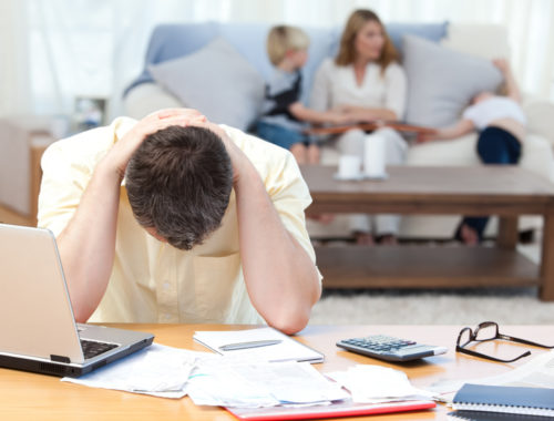 A man with his head in his hands calculating his debt expenses at a table with a laptop, calculator, eyeglasses, and debt papers on it.