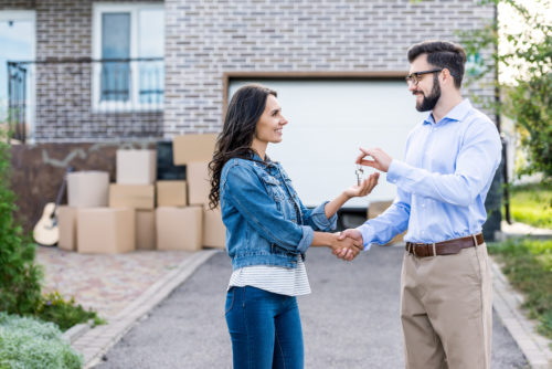 A man gives the keys to a first time home buyer in the driveway of the property with packing boxes in the background.