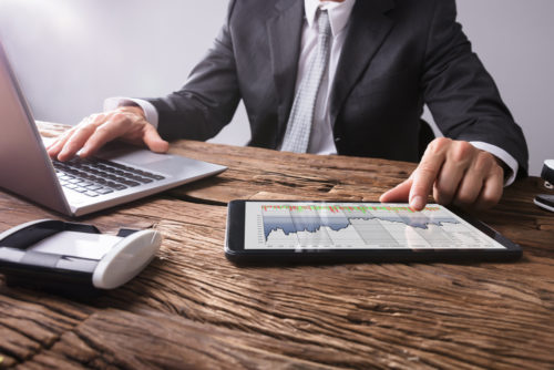 A stock broker calculates stock dividends on a computer and a tablet, showing a graph of stock figures.