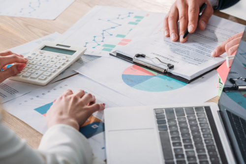 Two people calculating investments across from one another at a table that has papers, calculators, and laptops on top of it.